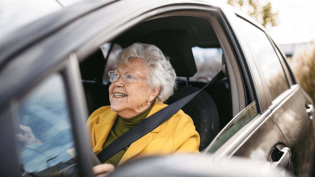 An elderly woman in a car, wearing a seatbelt, smiling and ready for her driving test.